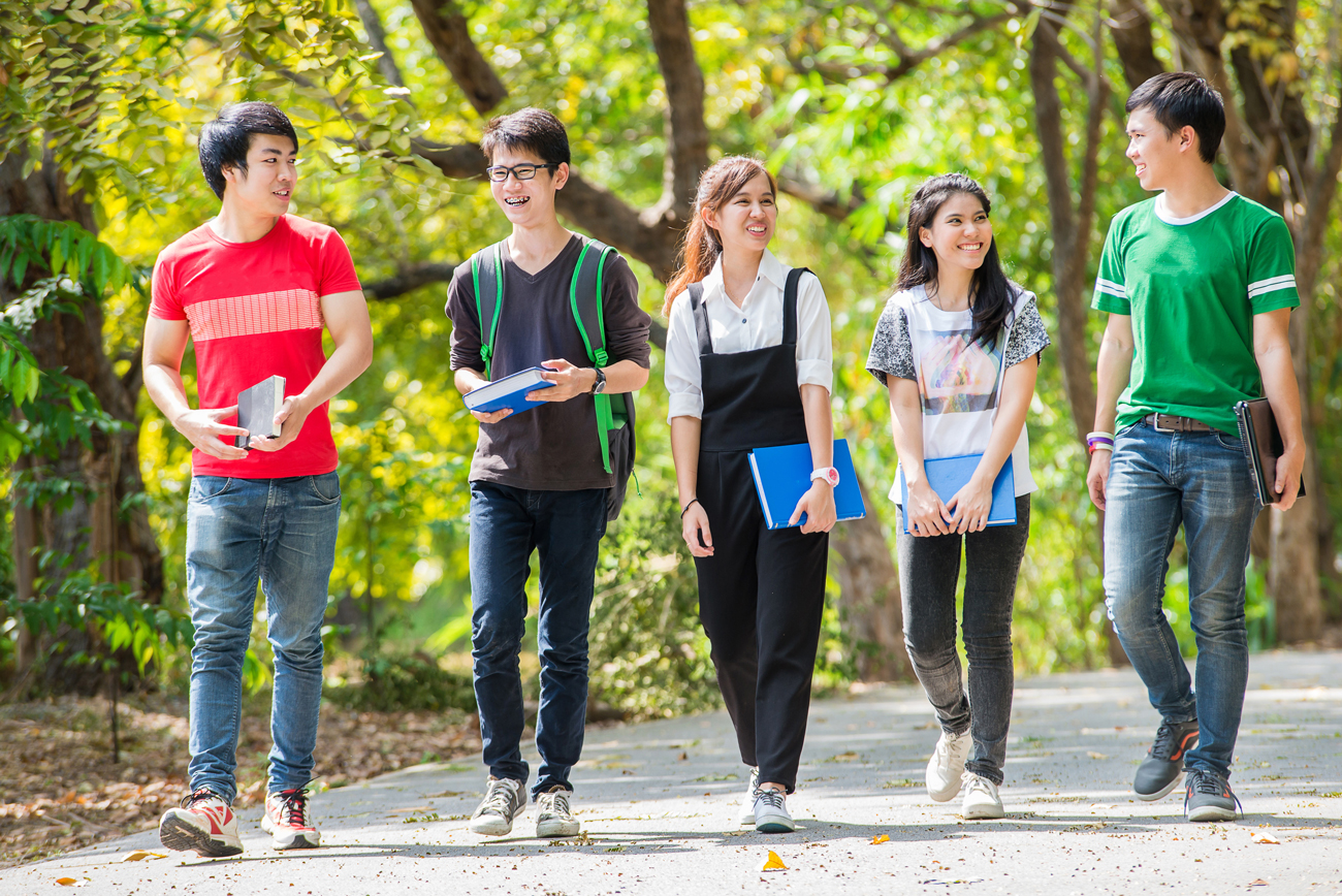 A group of five smiling young adults walking side-by-side on a paved path in a park while holding books and folders.