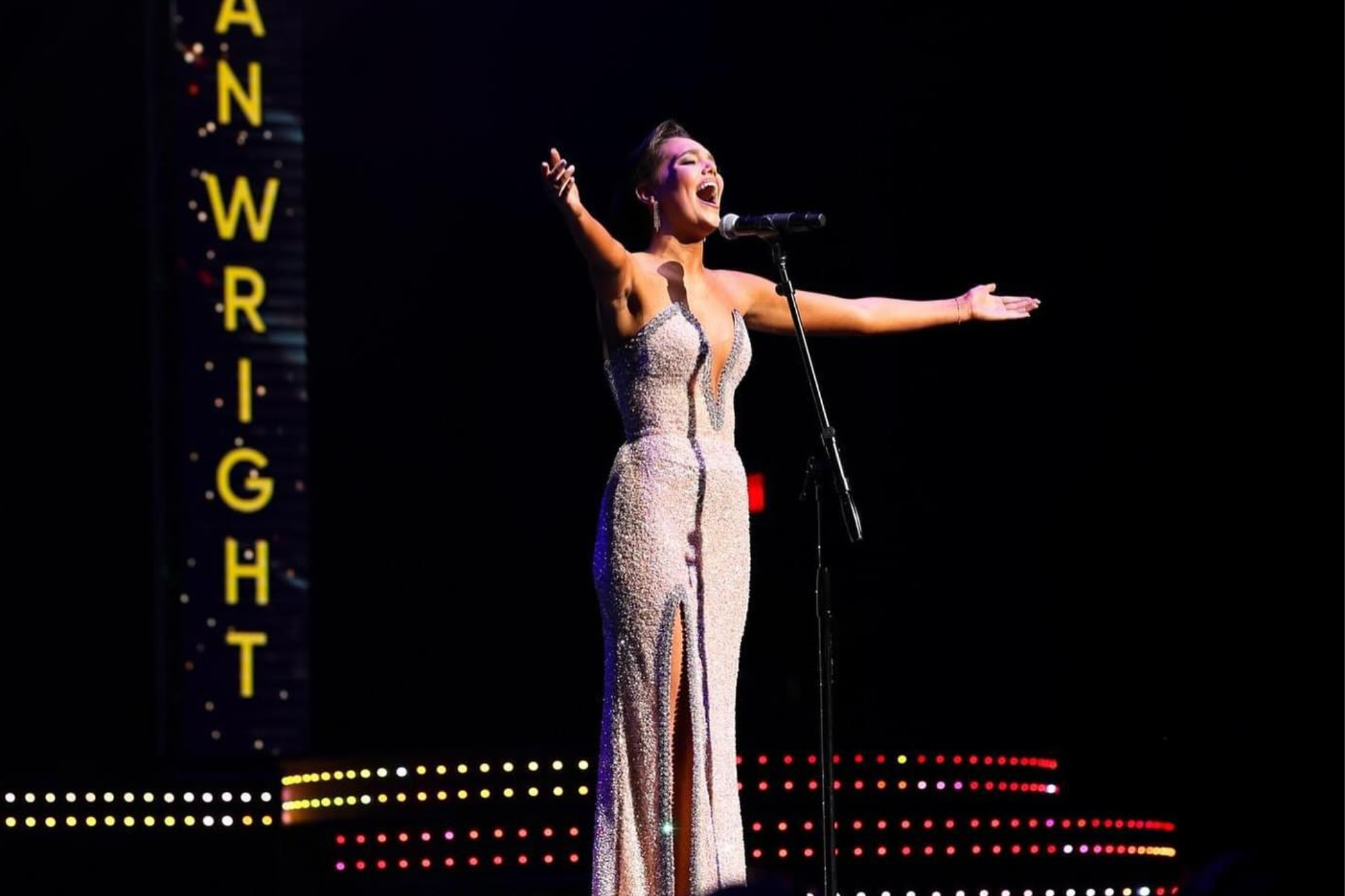 A woman in a sparkling, sleeveless evening gown with a high slit stands on a dark stage, singing into a microphone with her arms wide open.