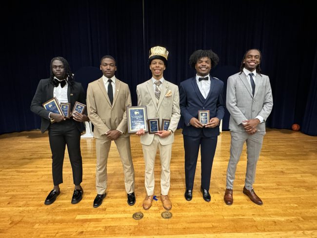 Five young men in suits pose together on a wooden stage holding award plaques; the central individual wears a gold crown.