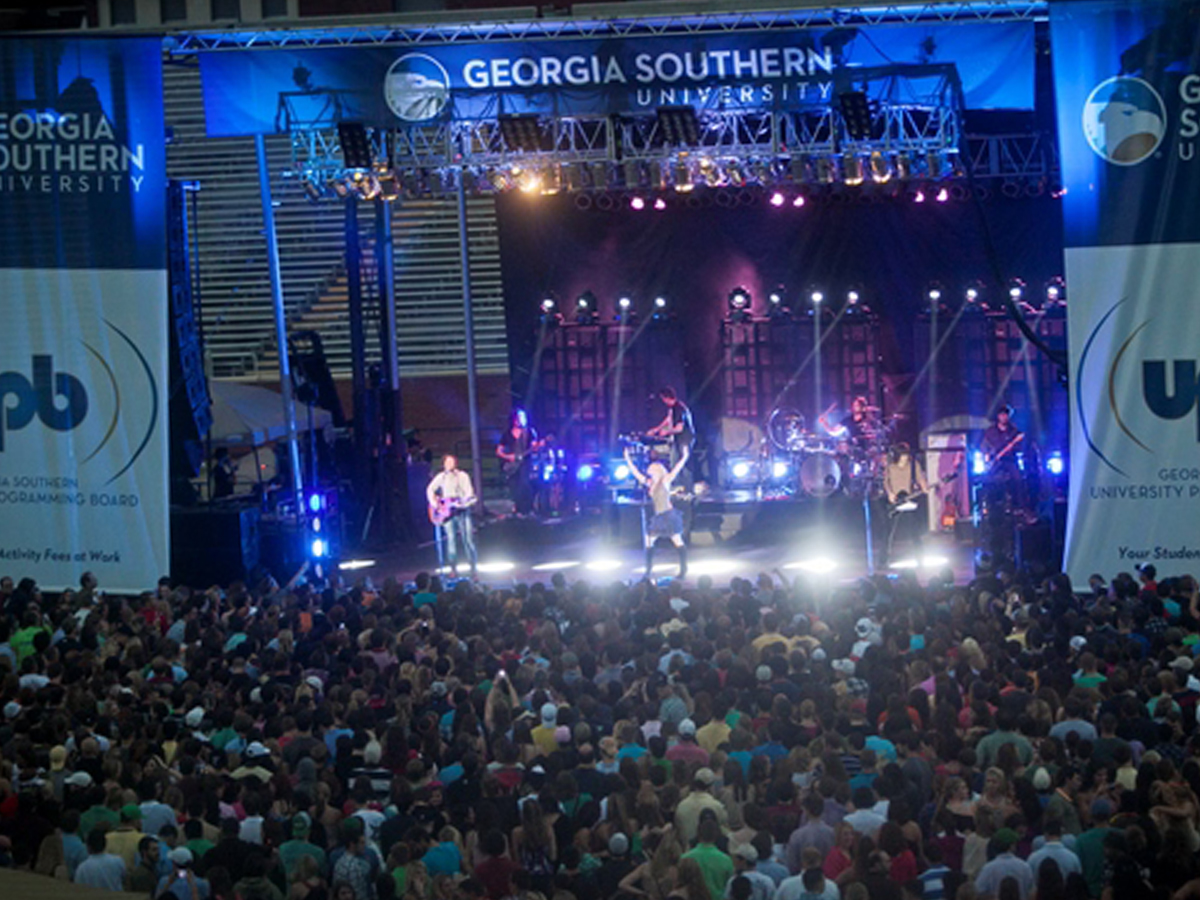 A rock band performs on a brightly lit outdoor stage under a Georgia Southern University banner for a large, packed crowd of concertgoers.