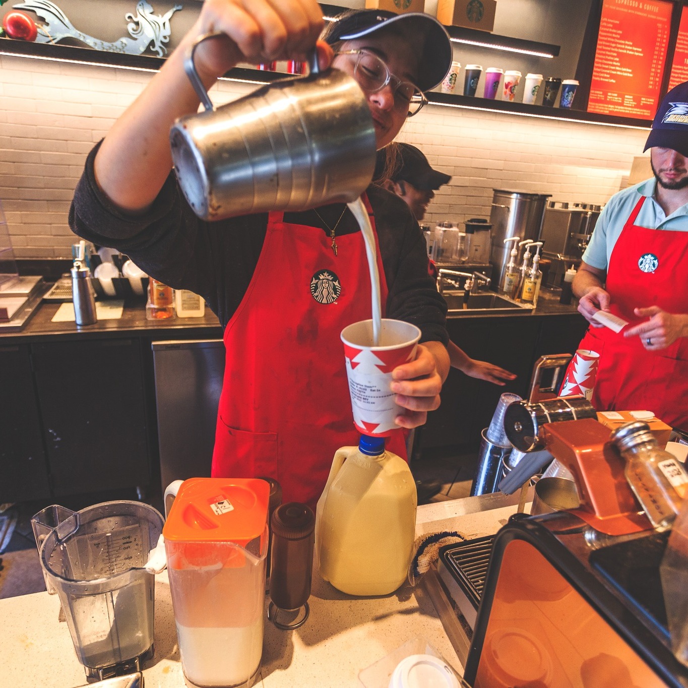 A barista wearing a red apron pours steamed milk from a metal pitcher into a paper coffee cup behind a busy Starbucks counter.
