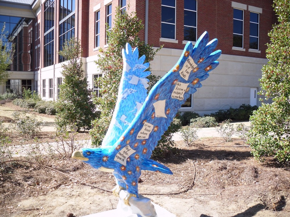A painted blue eagle statue decorated with stars, clouds, and quotes about knowledge stands on a university campus lawn.