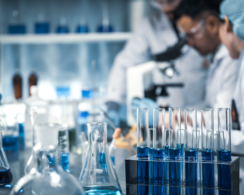 A row of glass test tubes filled with blue liquid stands in a rack on a laboratory counter, with scientists working out of focus in the background.