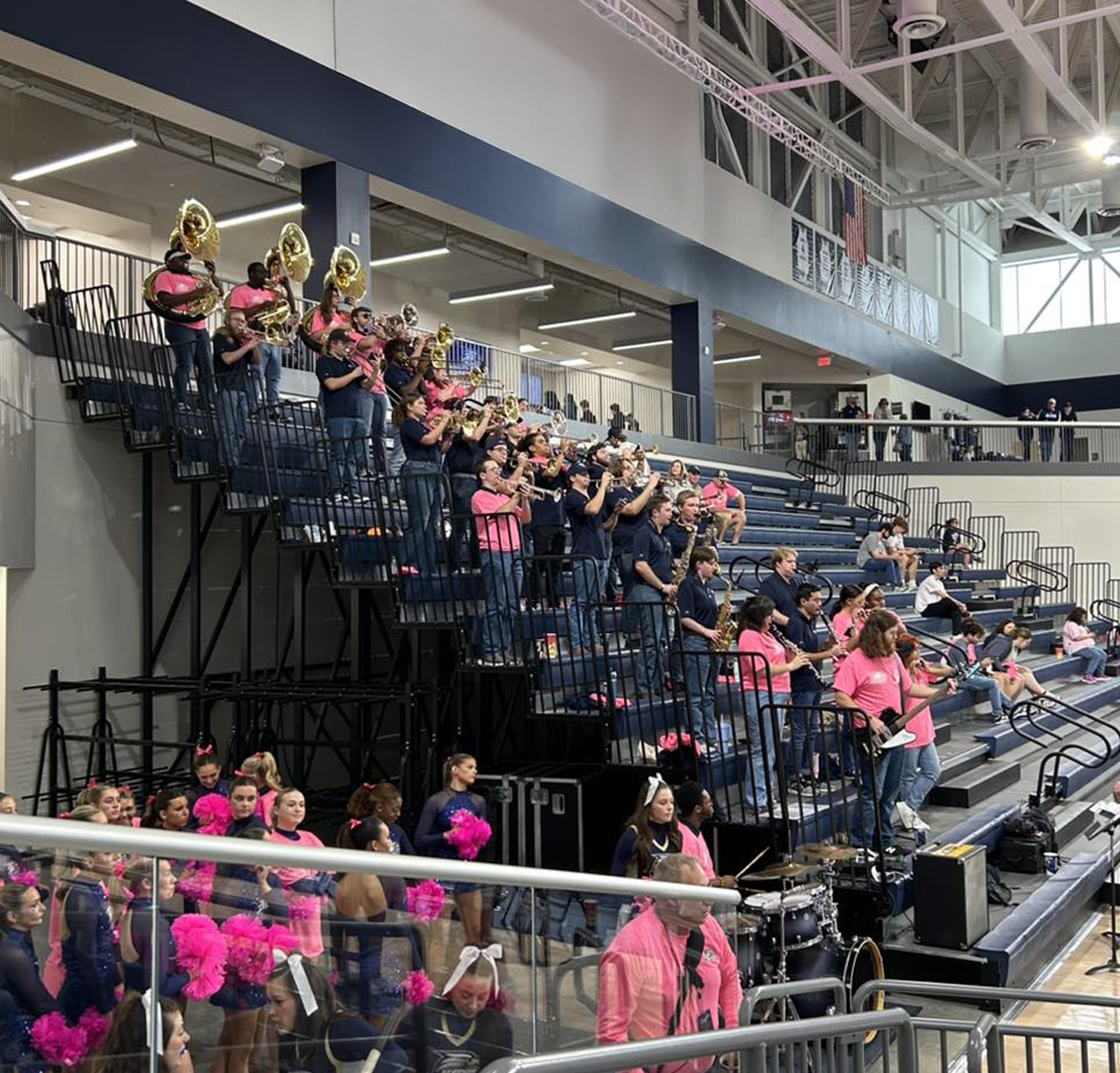 A pep band wearing pink and blue shirts performs with brass, woodwind, and percussion instruments from the bleachers inside a university gymnasium.