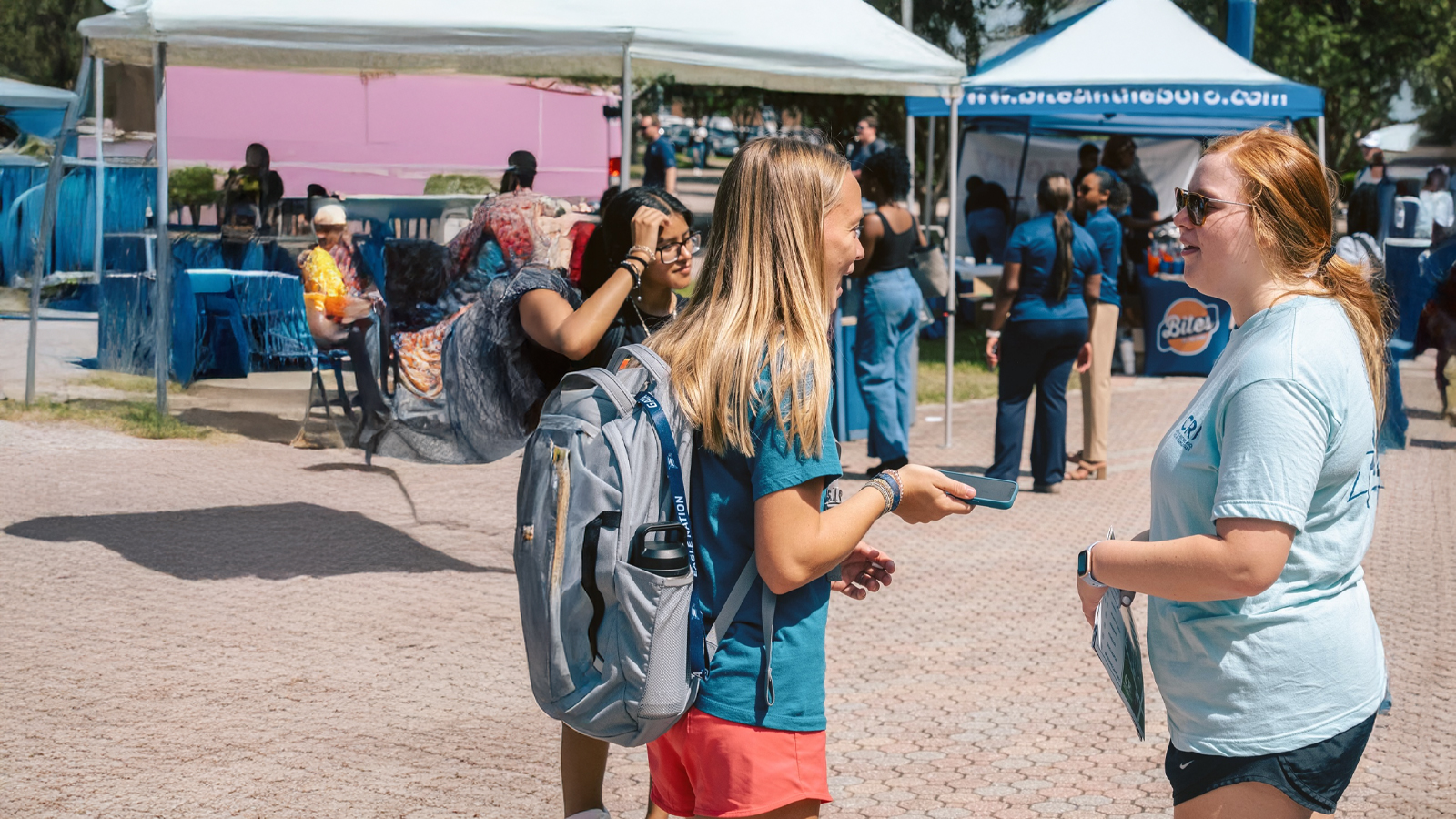 A lively outdoor student fair or campus event where a female student wearing a backpack chats animatedly with a representative at an information table.