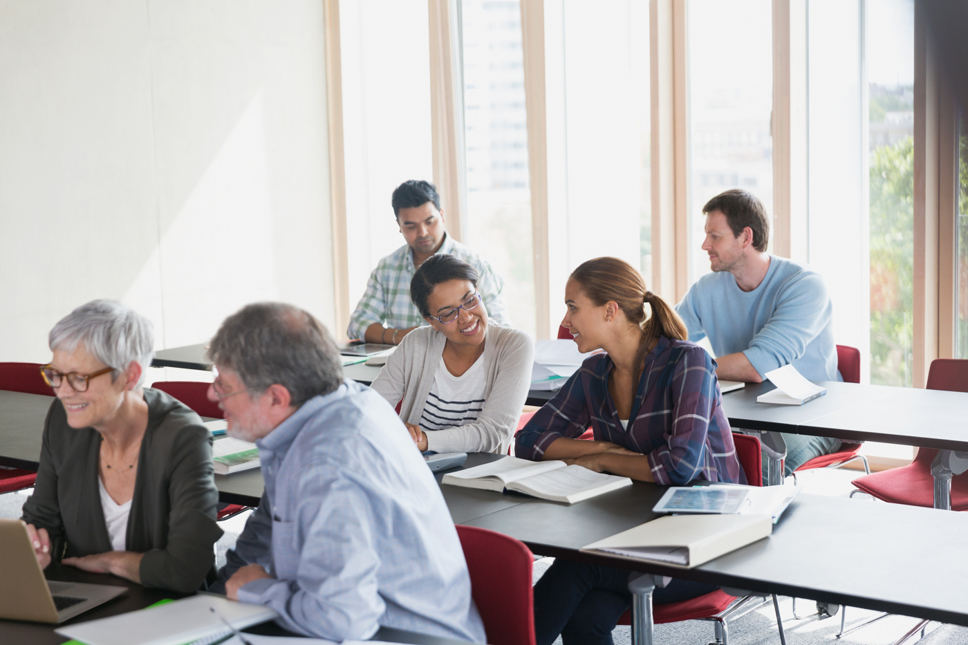 A diverse group of adult students sits at rows of desks interacting, talking, and looking over materials during a lecture or seminar.