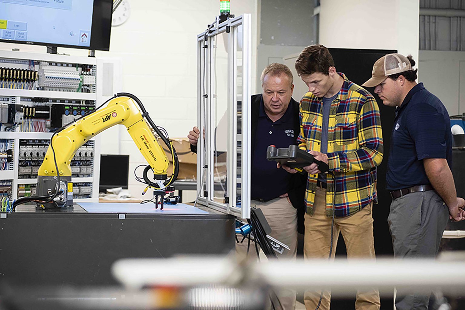 An instructor guides two students as they learn to program a yellow robotic arm using a control pendant in an advanced manufacturing lab.
