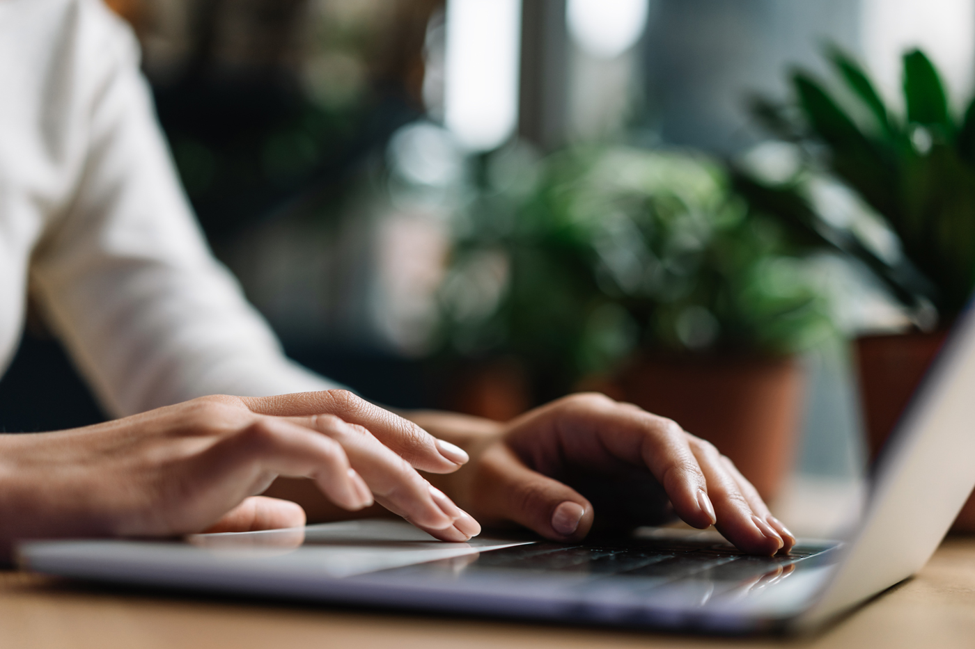 A close-up shot of a person's hands typing on a laptop keyboard at a wooden desk with blurred potted plants in the background.