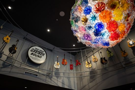A vibrant, multicolored blown-glass chandelier hangs from a dark ceiling above a display of vintage Gretsch electric guitars mounted on a curved gallery wall.