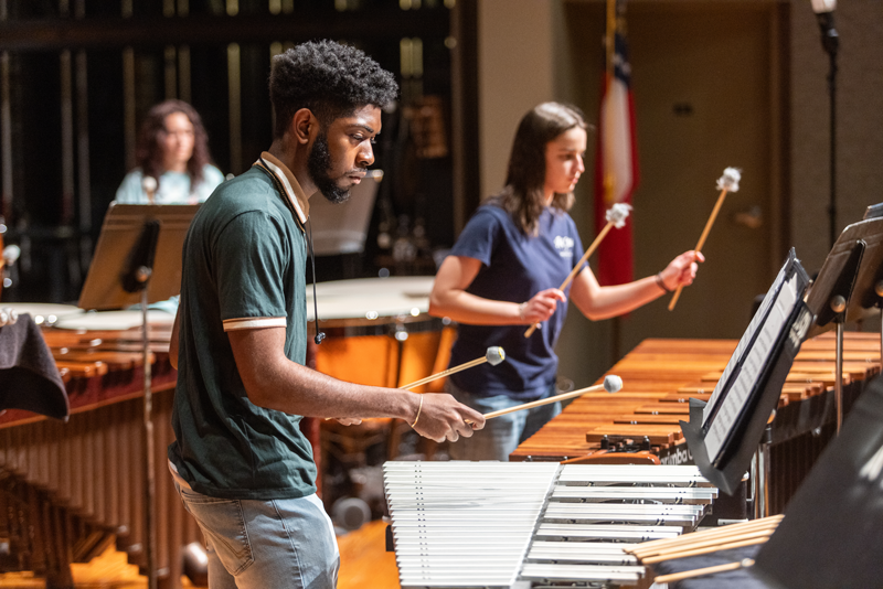 A close-up shot of a male student playing a vibraphone alongside a female student playing a marimba during a percussion ensemble rehearsal.