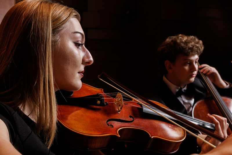 A close-up profile shot of a woman playing the viola alongside a man playing the cello in the background during an orchestral performance.