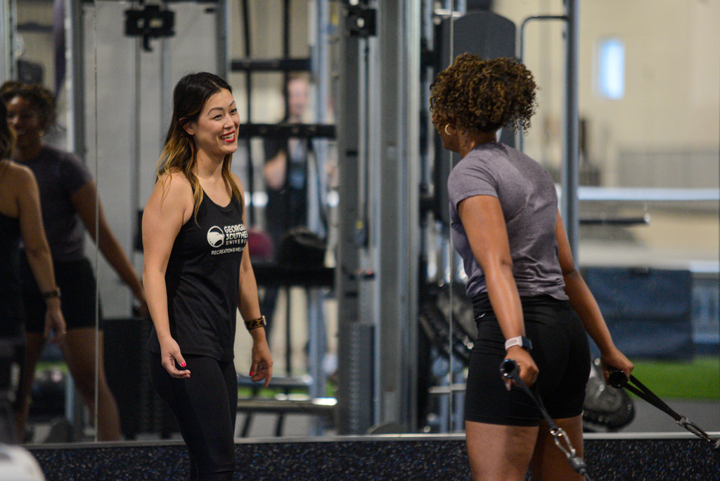 A fitness trainer in a black tank top smiles and talks with a woman who is performing a resistance band workout in front of a mirror at a gym.