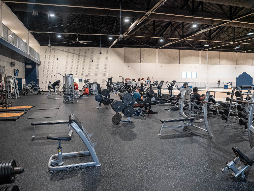 A wide shot of a spacious, well-equipped university recreation center weight room and gym floor filled with various weight machines, barbells, and cardio equipment as people exercise.