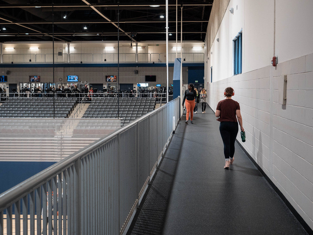 People walking and exercising on an elevated indoor track overlooking a large university gymnasium and a cardio fitness area with rows of treadmills.