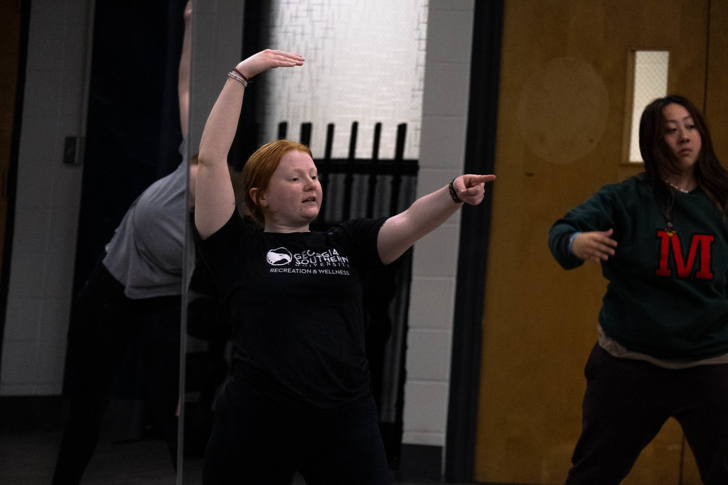 A dance instructor in a black Georgia Southern University recreation shirt leads a class with one arm raised overhead and the other pointing forward.