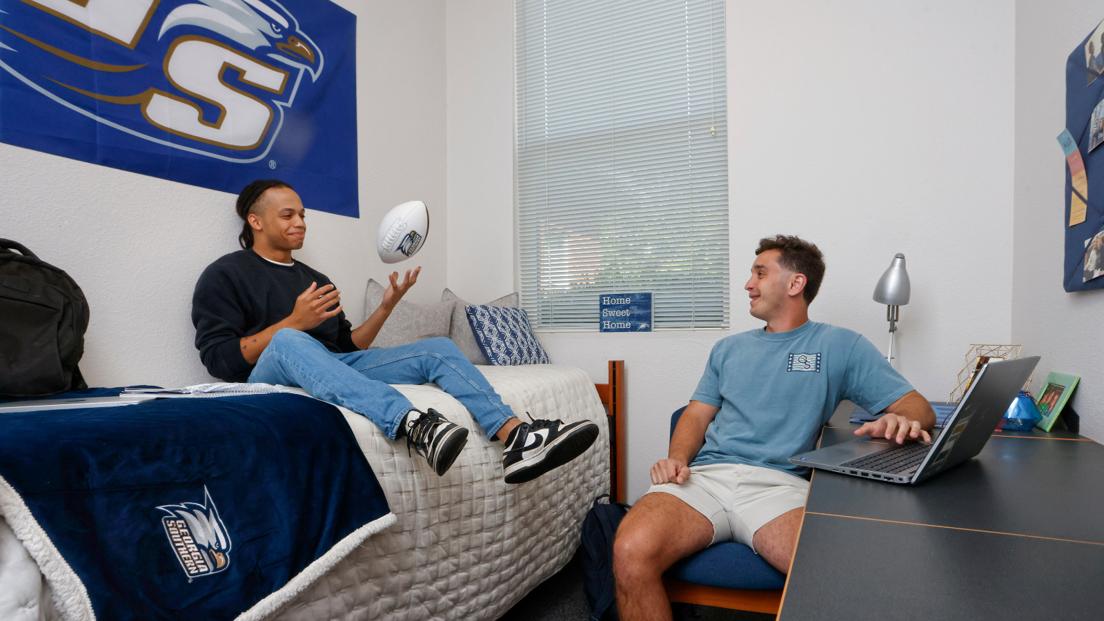Two men in a dorm room: one sits on a bed tossing a rugby ball while the other sits at a desk with a laptop, smiling at him.