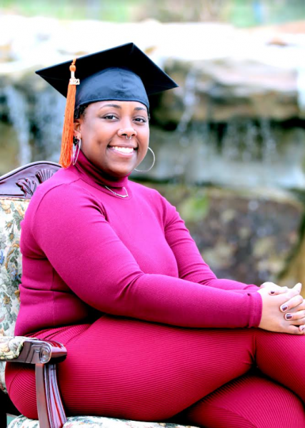 Valesia Davis sits in in front of small waterfall wearing bright pink attire and a graduation cap.