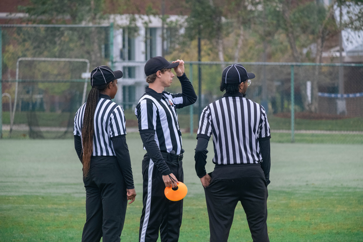 Three sports referees wearing black-and-white striped shirts and black caps stand together on an outdoor green athletic field while observing a game.