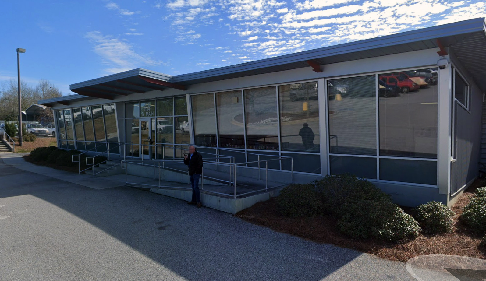 Exterior daytime shot of a modern single-story commercial building with an asymmetrical roof line, large glass windows, an accessible entrance ramp, and a man standing outside.