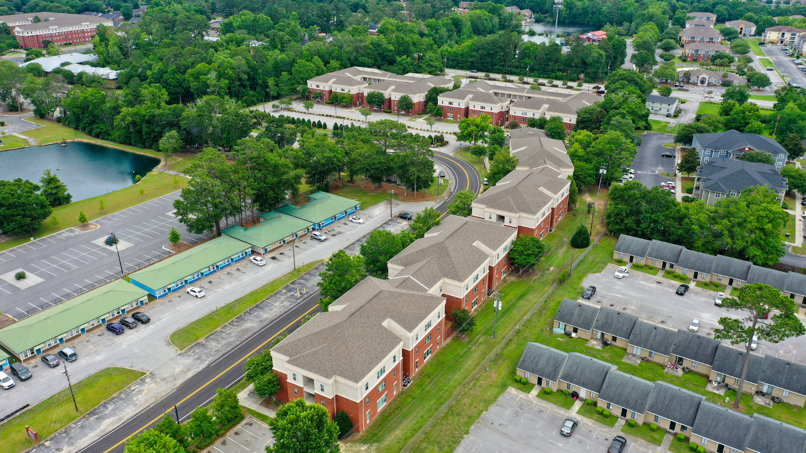 A third copy of the high-angle panoramic view overlooking the campus residential complex, showing the buildings with green metal roofs, brick facades, and winding courtyard walkways surrounded by green trees.