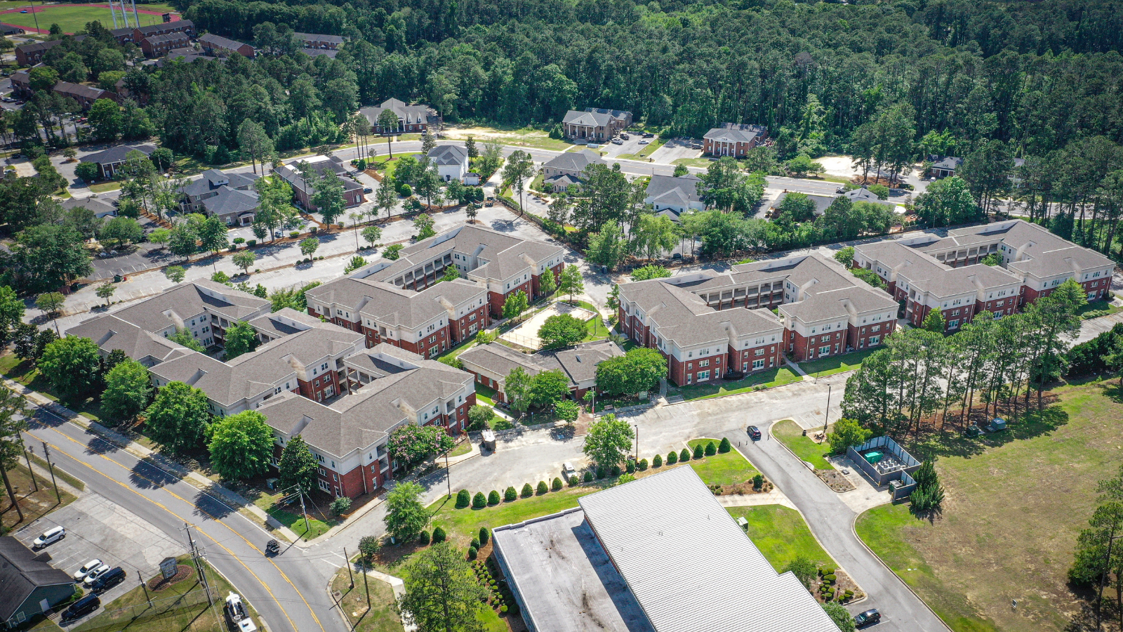 A wide, high-angle aerial drone photograph showcasing a massive multi-building campus apartment complex. The buildings feature a mix of brick and light-toned upper levels with gray shingled roofs, arranged in U-shaped courtyards. The complex is flanked by a roadway with yellow lines, paved parking lots, manicured lawns, and clusters of tall green pine trees.