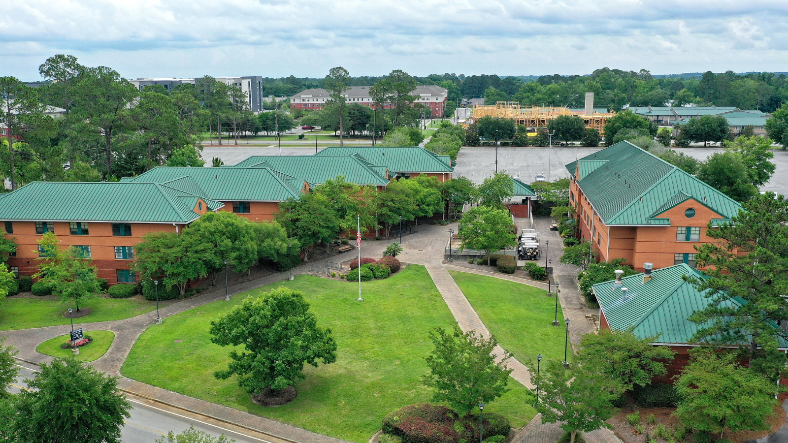 An aerial, high-angle panoramic view overlooking a campus residential complex. The buildings feature distinct green metal roofs, brick facades, and central courtyard spaces connected by concrete walkways and manicured lawns, with construction visible in the distant background.