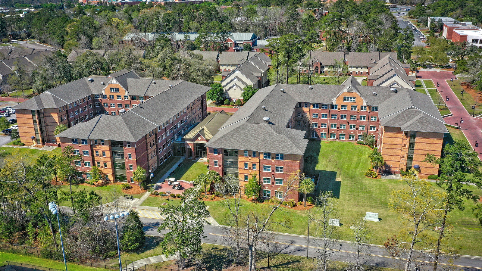 A high-angle aerial drone photograph showcasing another distinct campus housing facility. The complex features two large, multi-story brick residential halls with architectural gables and gray shingled roofs, arranged around an open grassy lawn with paved brick walkways and scattered trees.