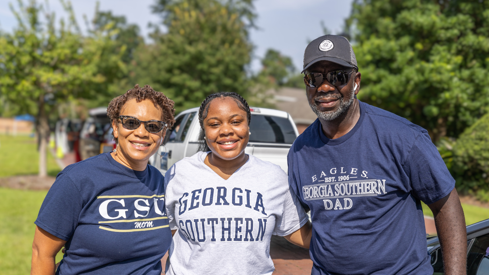 Picture in sunny, outdoor family portrait featuring a smiling female college student flanked by her parents. They are all wearing Georgia Southern University gear—the mother wears a dark blue 