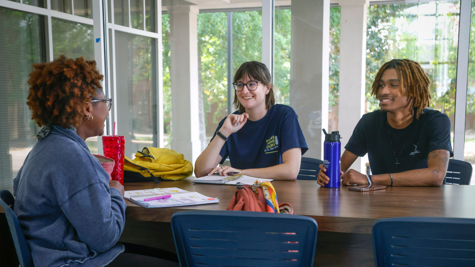 A bright, candid photo of three college students sitting together at an outdoor or covered campus table, smiling and chatting while studying with notebooks, water bottles, and backpacks open in front of them.