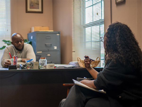 An over-the-shoulder view of a woman with curly hair holding a pen and taking notes during a meeting with a man sitting at an office desk.