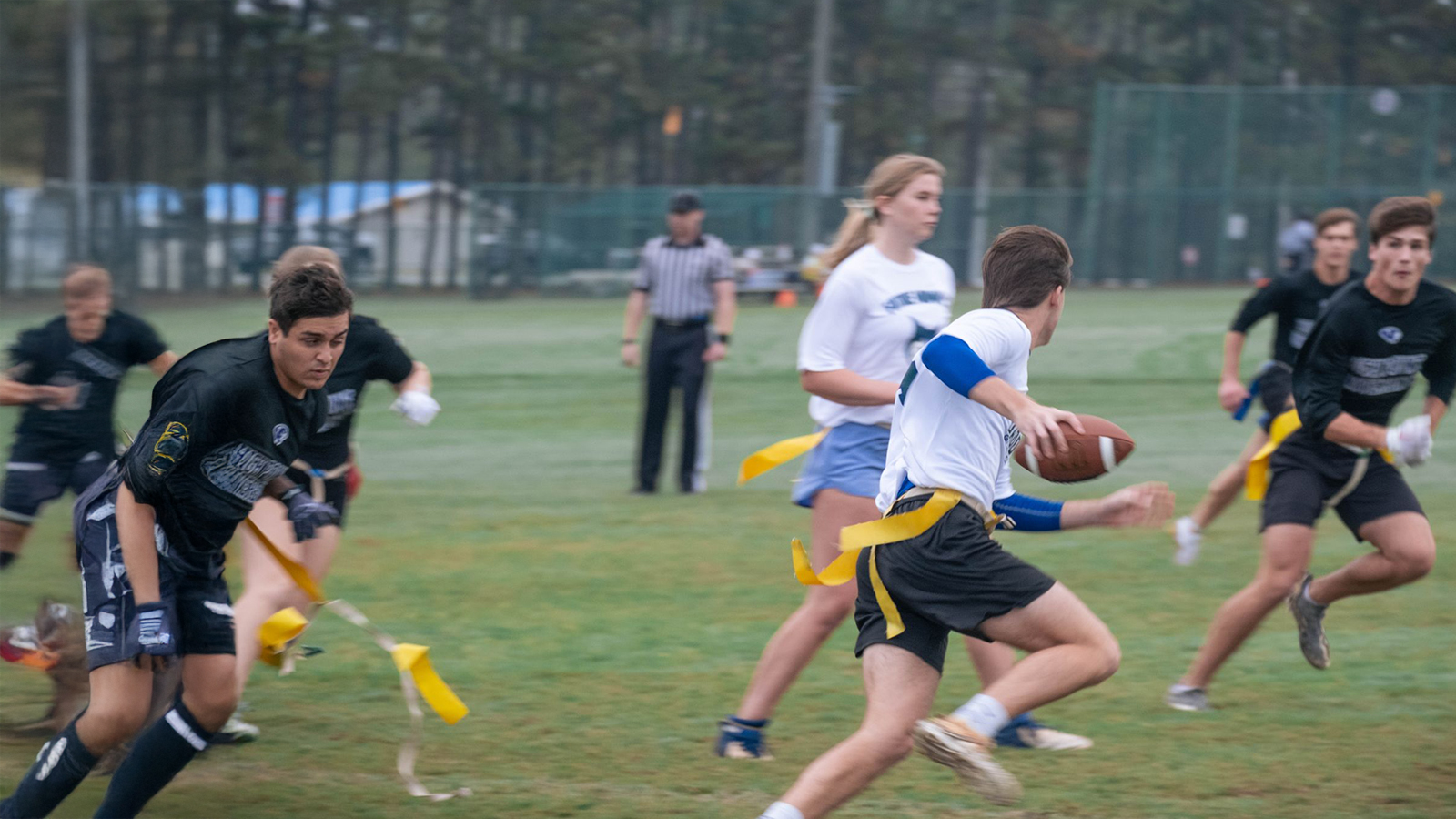 Dynamic action shot capturing an intramural flag football game on a green campus recreation field. The image shows a quarterback in a white shirt and blue undersleeves preparing to throw a pass while scanning the field, with oncoming defenders in black jerseys trying to pull his yellow flags.