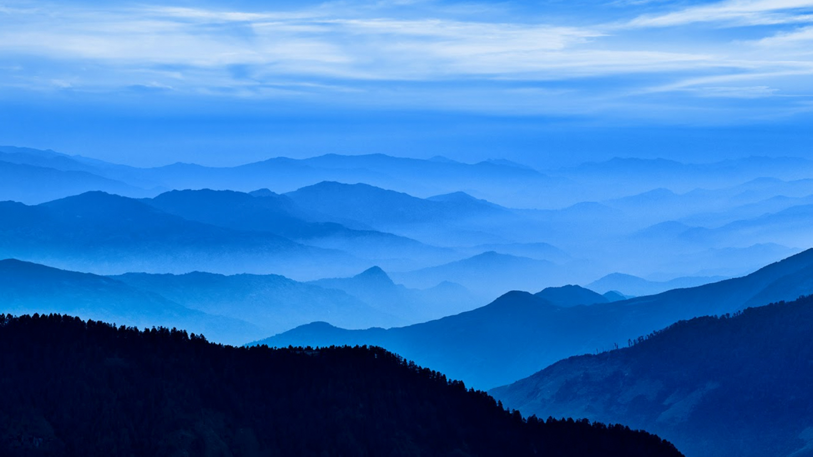 A breathtaking, monochrome-blue panoramic view of rolling mountain ranges fading into a misty horizon, creating a beautiful layered silhouette effect.