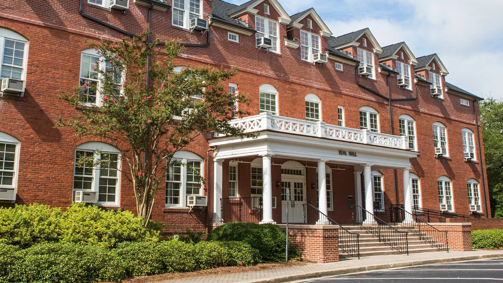 Exterior shot of the historic, multi-story brick Deal Hall, featuring white-pillared porch columns, classic balcony railings over the entrance stairs, and landscaped hedges.