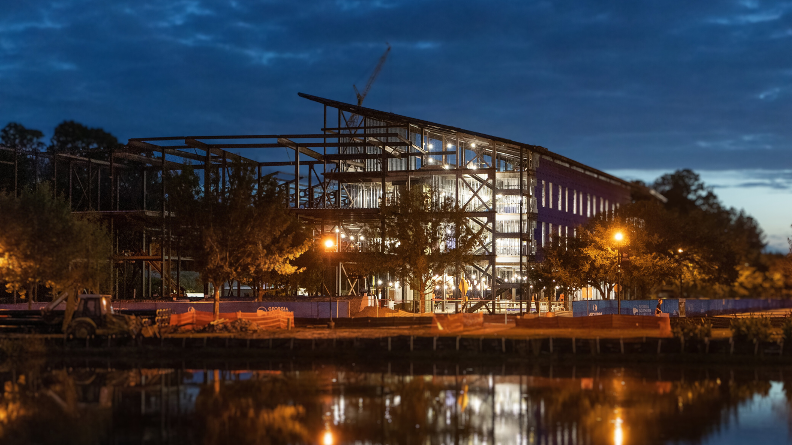 A nighttime view of a large university building under construction, illuminated by bright work lights with its structural steel framework reflecting on a body of water in the foreground.