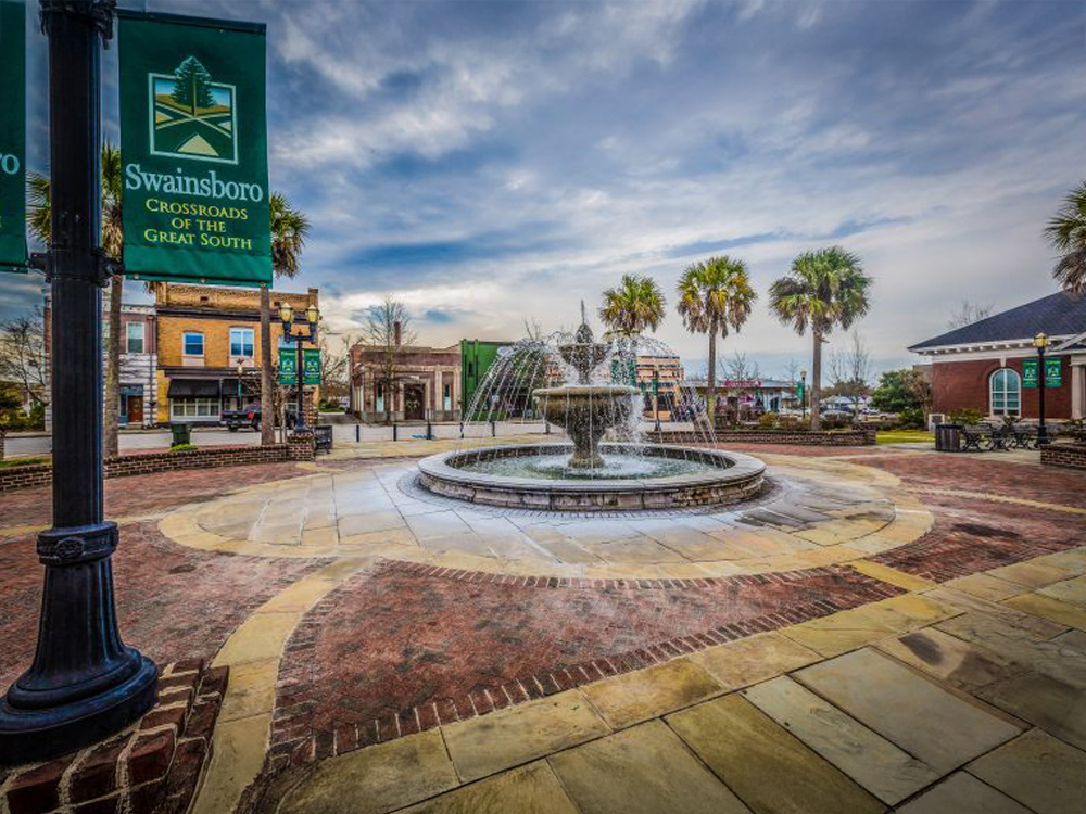 wide-angle photograph capturing the town square fountain in Swainsboro, Georgia. A prominent green banner on a lamppost reads 