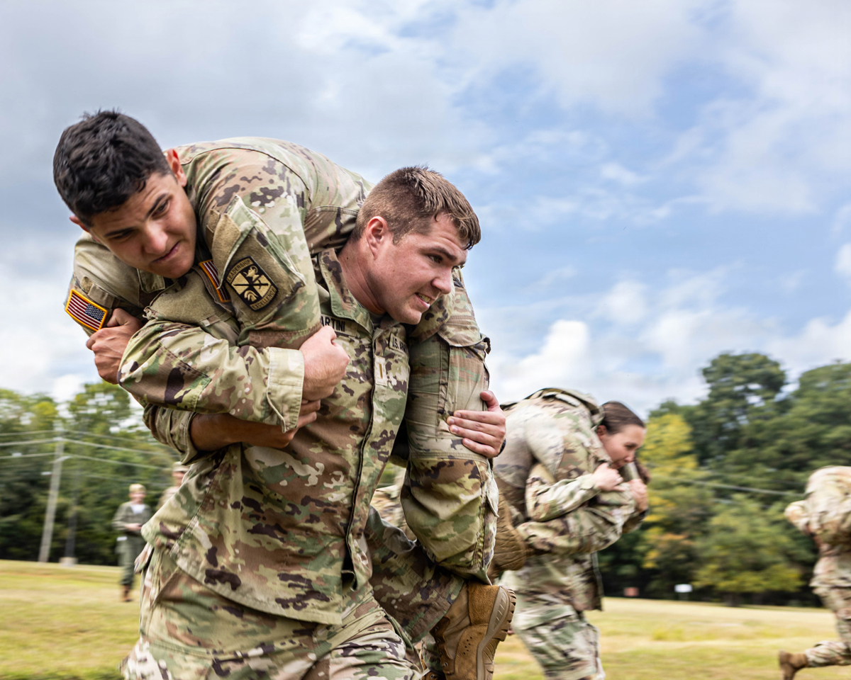 Two soldiers in camouflage uniforms perform a fireman's carry during a training exercise on a grassy field under a cloudy sky.