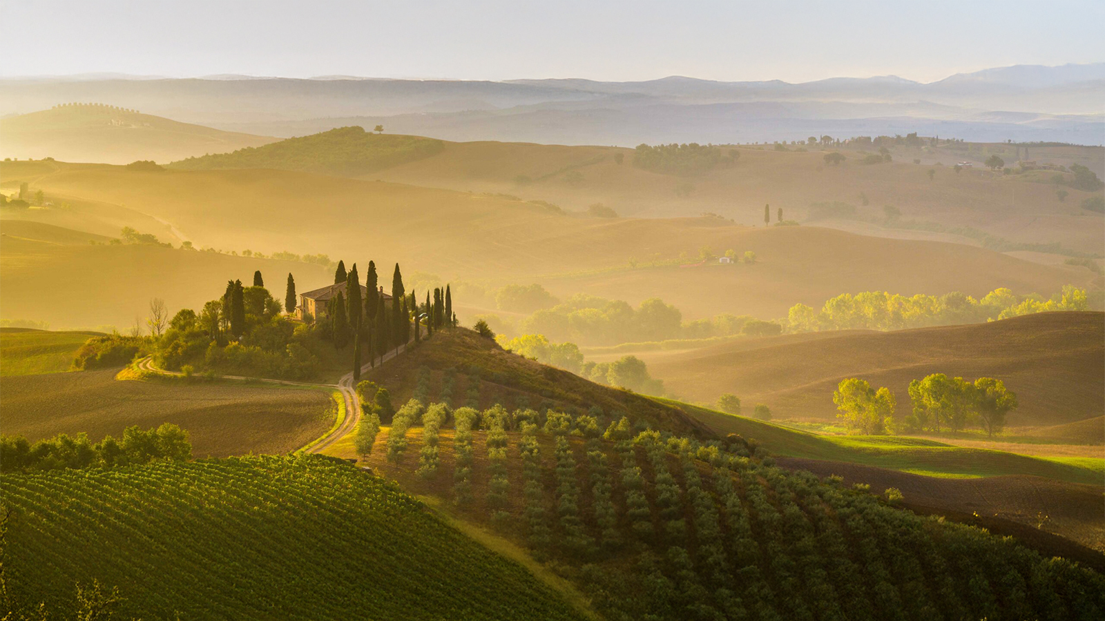 A serene villa sits atop a rolling Tuscan hillside at sunrise, surrounded by slender cypress trees and misty, golden fields that stretch toward distant mountains.