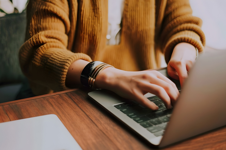 A person wearing a mustard-yellow ribbed sweater and stacked bracelets types on a laptop keyboard at a wooden desk.