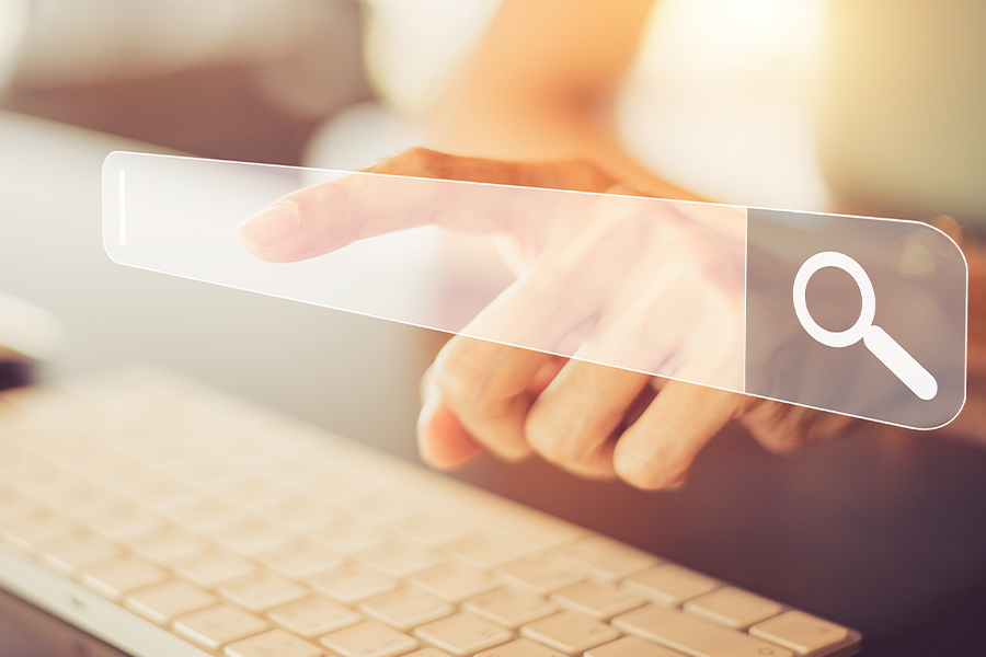 A hand reaches out to touch a translucent, digital search bar overlaying a blurred background of a desk and computer keyboard.