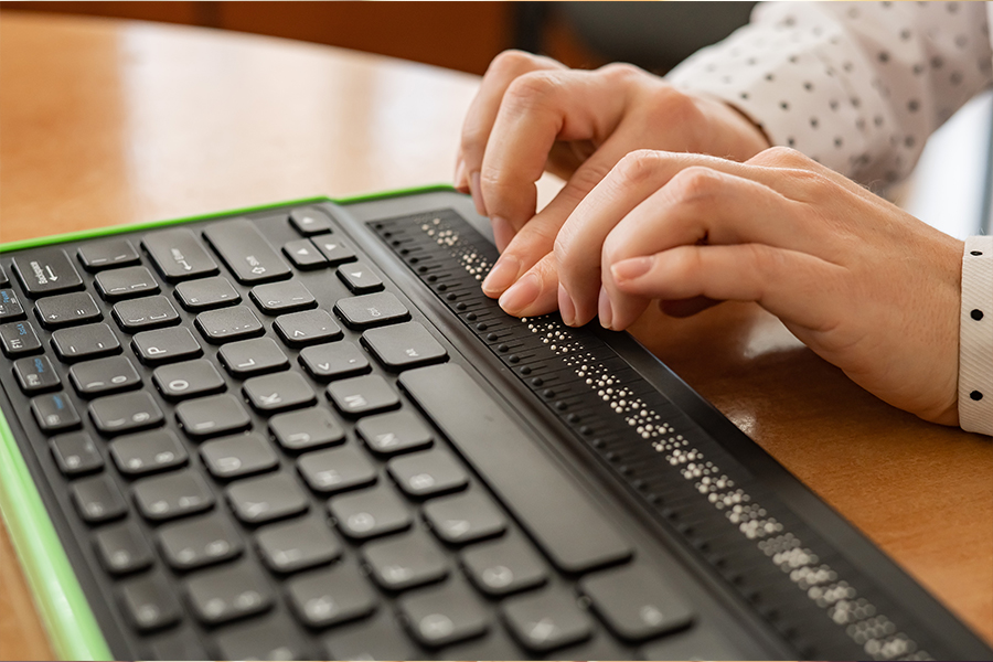 A close-up shows a person's hands using a refreshable Braille display integrated into a computer keyboard to read text through touch.
