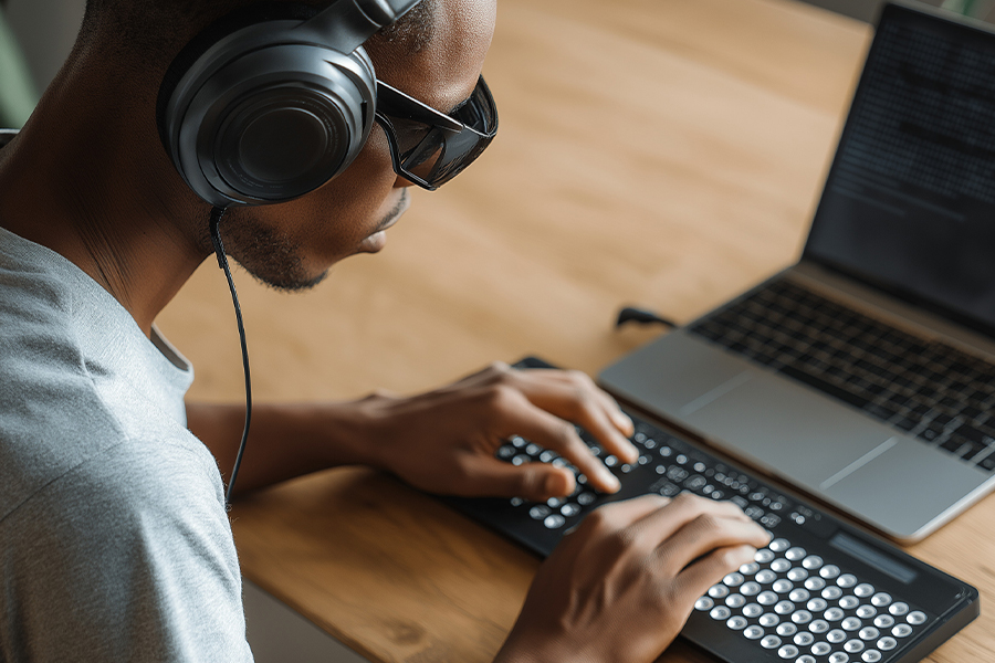 A person wearing headphones and dark glasses works at a desk, using a specialized keyboard with large, tactile buttons connected to a laptop.
