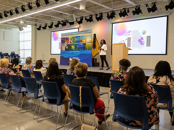 A speaker in a white blazer presents to an audience about health logistics and analytics, with digital screens displaying maps and project data on stage.
