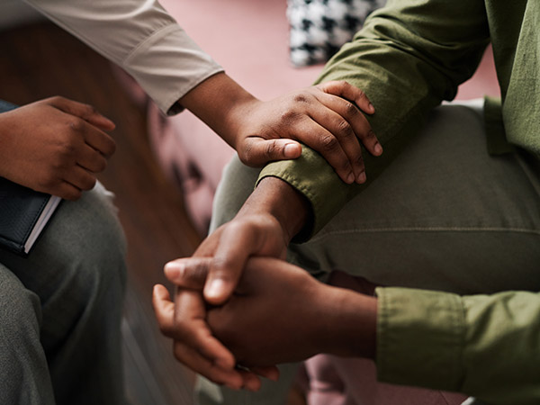 A close-up, high-angle shot shows one person's hands resting supportively on the arm and hand of another person during a comforting conversation.