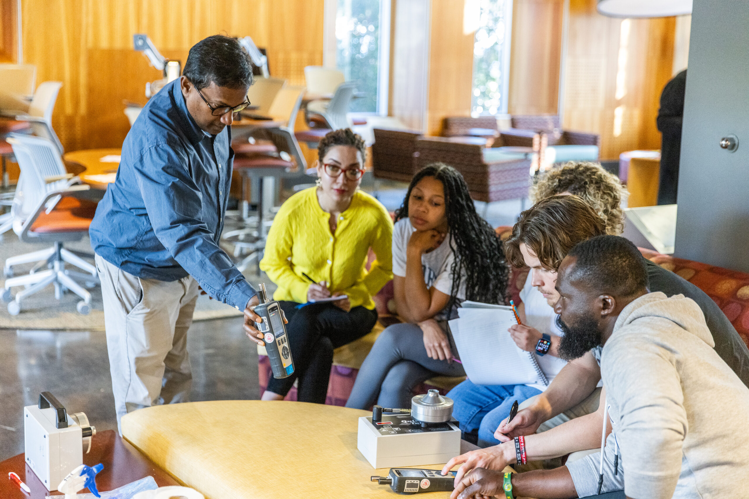 An instructor demonstrates the use of a handheld environmental monitoring device to a group of five attentive students in a brightly lit, modern lounge area.