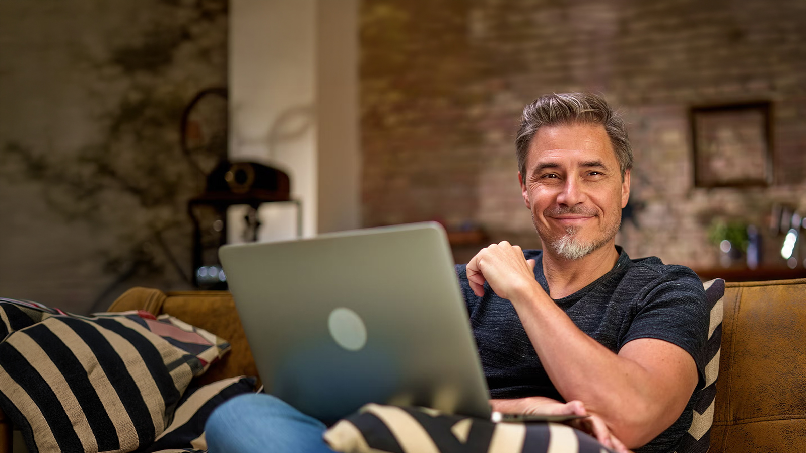 A middle-aged man with salt-and-pepper hair and a goatee smiles warmly while sitting on a couch with a laptop in a cozy, dimly lit room with exposed brick.