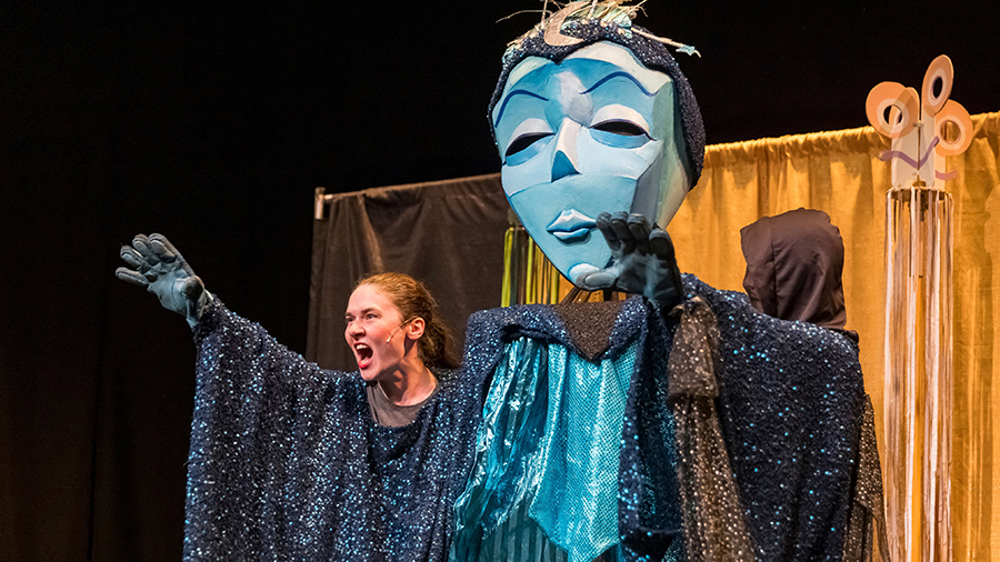 Theatrical performance featuring a large blue masked puppet figure in a glittery cloak, with a performer in a sparkling cape gesturing dramatically on stage
