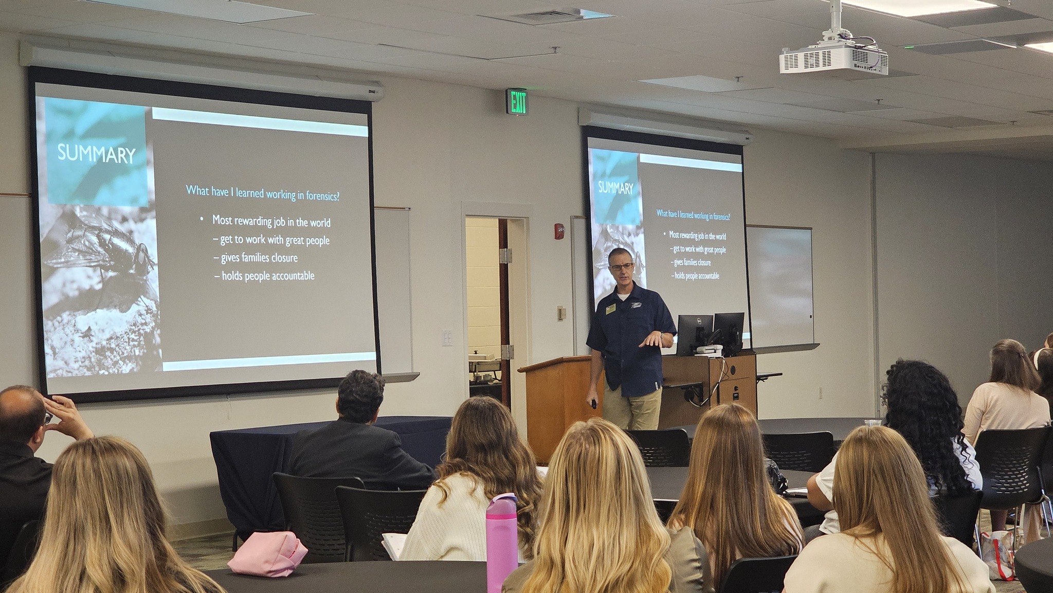 An instructor stands in front of a group of people at a conference. The attendees are seated in chairs grouped at tables. The presentation screen at the front of the room describes forensics as the best job in the world.