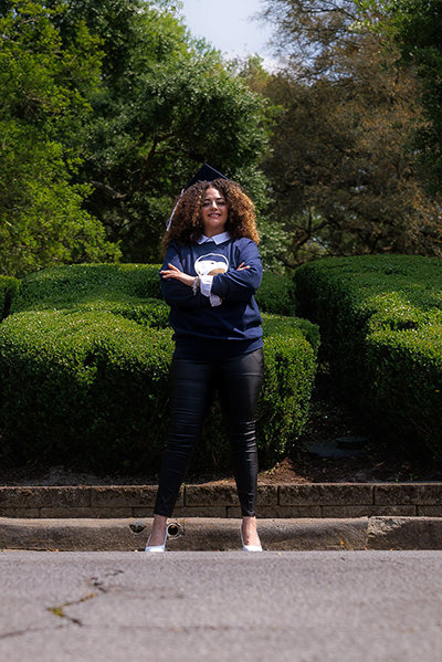Graduate in a GSU tshirt standing with arms crossed on a path, in a park with trimmed hedges behind her.