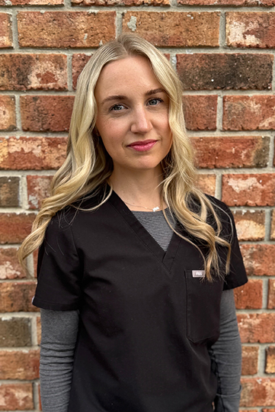 Woman with long blonde hair wearing black scrubs over a gray long-sleeve shirt, standing in front of a brick wall.