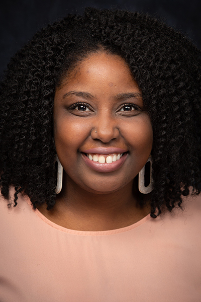 Close-up portrait of a smiling Black woman with curly hair, wearing a peach top and rectangular earrings.